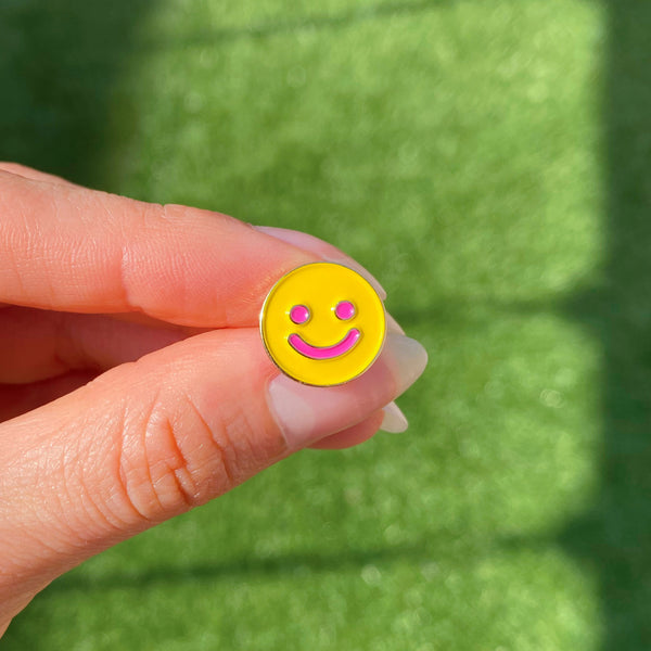 hand holding a single smiley earring (yellow round smiley with pink eyes and mouth)
